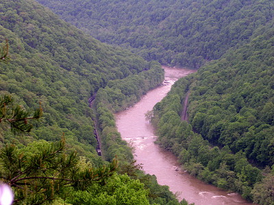 [Tubulent, muddy river snaking through the gorge. A train track is visible on the left bank. The steep hillsides on both sides of the river are blanketed with leaf-bearing trees.]